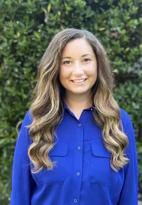 A woman with long hair standing in front of bushes.